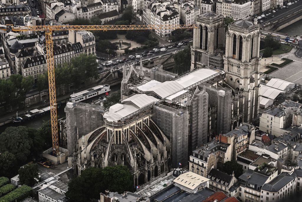 An aerial view of the exterior of the cathedral, its damaged roof mostly covered by white tarp. An enormous crane looms over the building