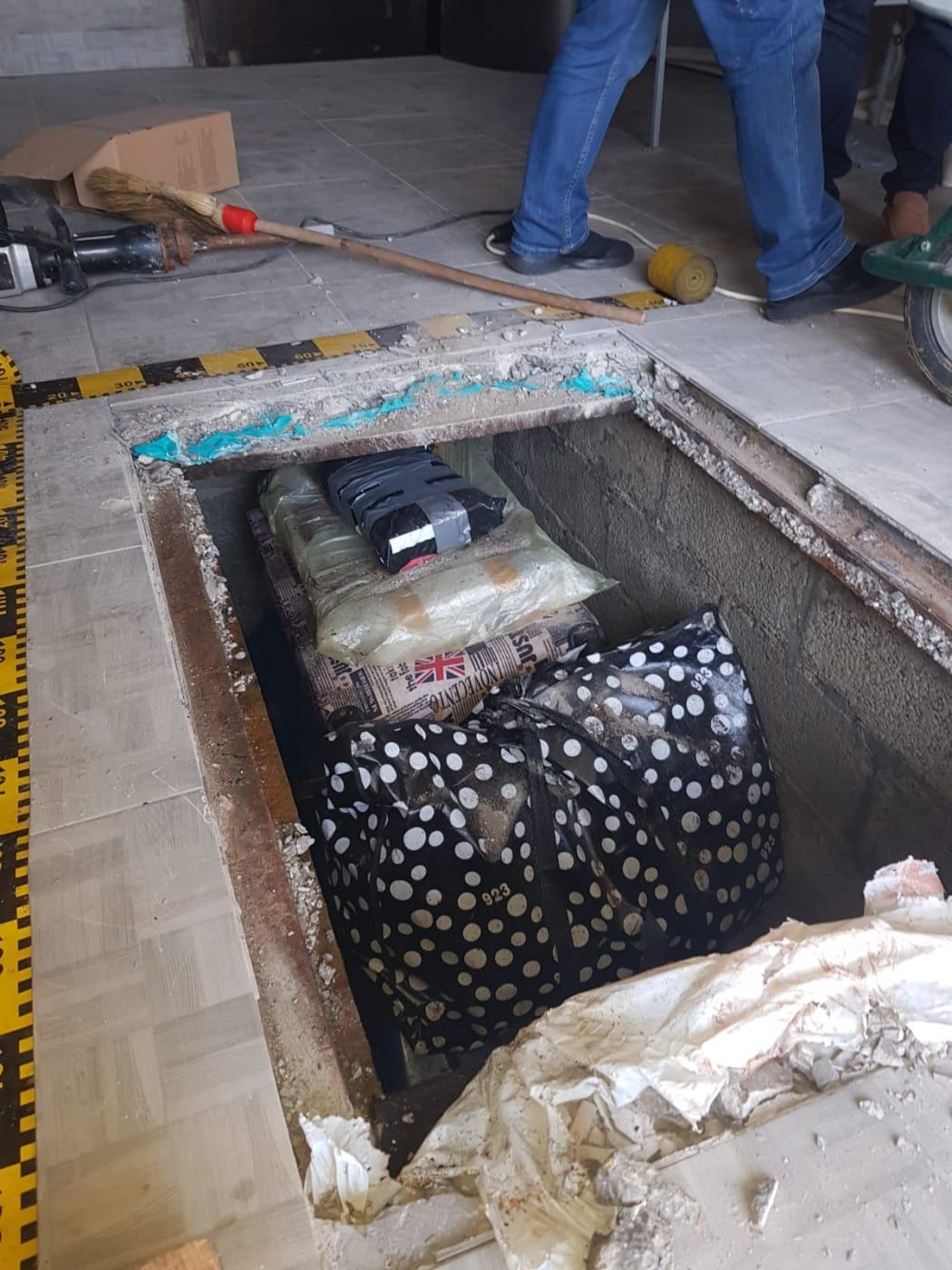 A view of a tiled floor, with a person's legs in blue jeans standing nearby. A large square has been cut into the floor, surrounded by crime scnee tape, and a big polka-dotted bag and many other plastic bags holding the books are stacked in the hole