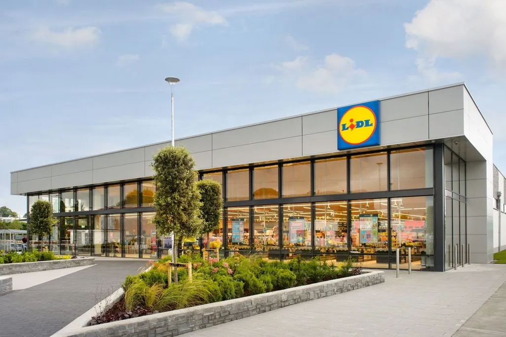 An image of a brightly lit new Lidl store; a large square building with the store's logo, a blue square with a yellow circle and blue and red block text that reads LIDL; surrounded by pavement and a parking lot