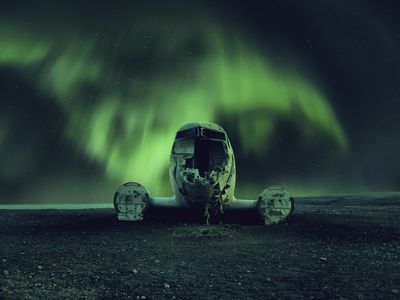 The northern lights cast an eerie glow upon an abandoned, wrecked airplane.

