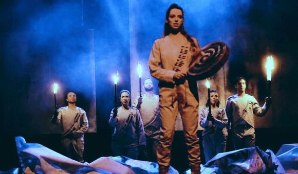 A woman stands on stage, holding a hand drum and a beater stick. Five actors behind her hold up torches. They all wearing matching white outfits with mock bandoliers. The stage is foggy and lit blue.