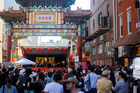 A crowd gathers on a street, where a stage is set up under an elaborate, decorative Chinese-style archway.