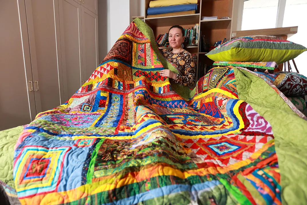 A woman holds up a large, brightly colored patterened quilt.