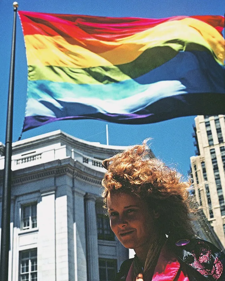 A woman with brown curly hair stands and smiles in front of a rainbow colored flag blowing in the wind in front of a stately white building and blue sky