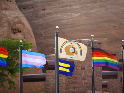 Various Pride flags (rainbow, transgender, equality, LGBTQ+ people of color, and love rainbow) displayed alongside the Navajo Nation flag in Window Rock, Arizona. (Photo by Pamela J. Peters (Diné)/Táchii’nii Photography, used with permission) 