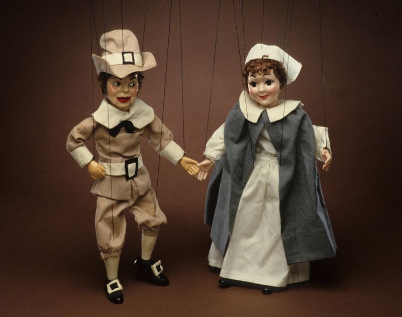 Two marionettes, a man and a woman, dressed in the clothes associated with Pilgrims, such as a men’s hat with a prominent buckle.