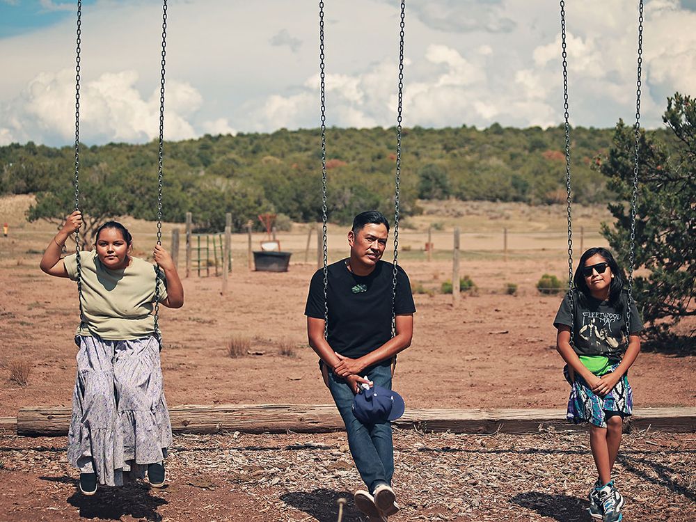 Two children and an adult sit on three swings in a dry landscape with trees in the background.