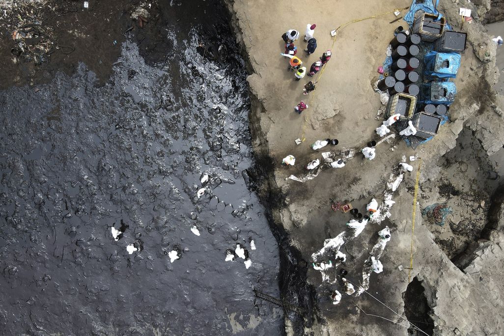 An aerial image of an oil spill off Peru's coast. The photo shows a contaminated beach smeared with black crude oil and workers in PPE trying to clean up the beach.