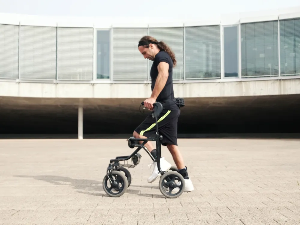 A man with long hair walks with the help of a walker on a concrete surface