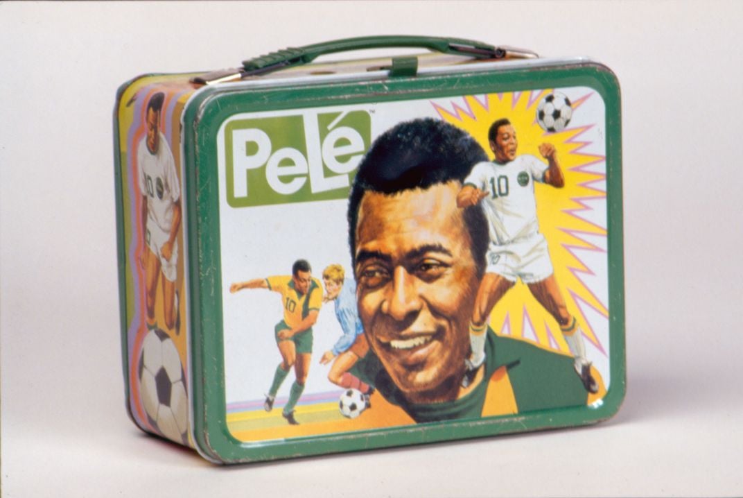 White and green children’s lunchbox. Three images of Pelé are displayed on the front cover. In order from left to right: Pelé plays soccer with a boy in the Brazil kit, portrait of Pelé wearing the Brazil jersey, and Pelé performs a soccer ball trick in t