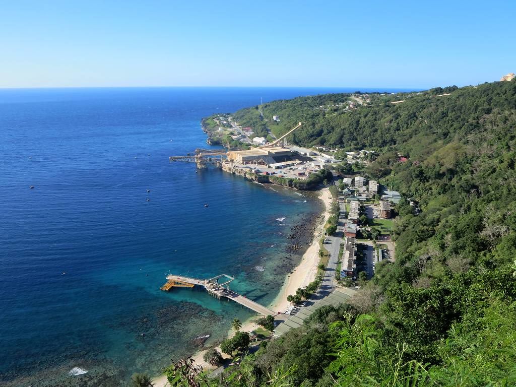 An image of a town on the coast of Christmas Island. The right side of the photo has forested hills with a town stretched along the coast. On the left is the bright blue ocean.