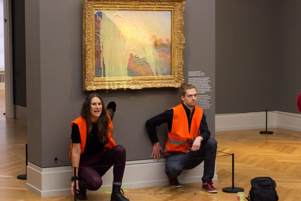 Two people with hands stuck to wall under painting smeared with potatoes