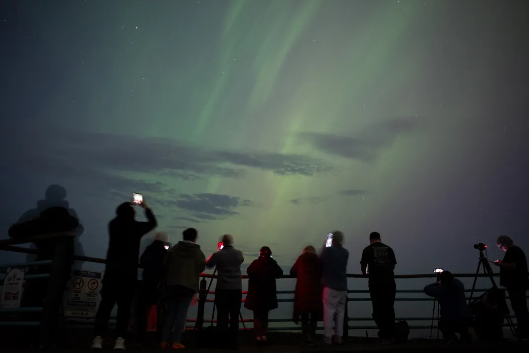 people stand along a railing taking pictures of the green-streaked sky with their phones and cameras