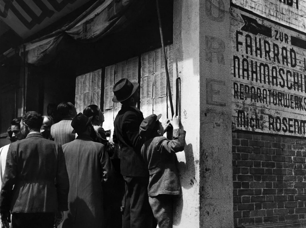 A group of men and some boys gather close to a posted list of names; on the side of a building, German writing is visible
