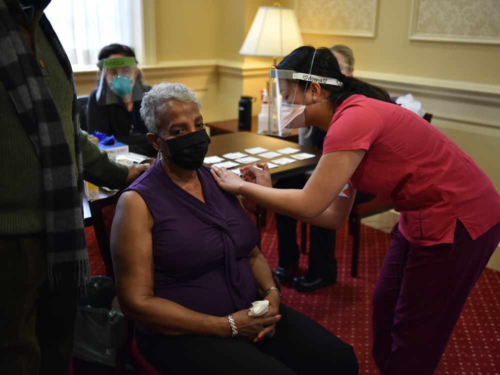 A woman sits in a chair and gets a vaccine shot from a woman wearing a face mask and shield