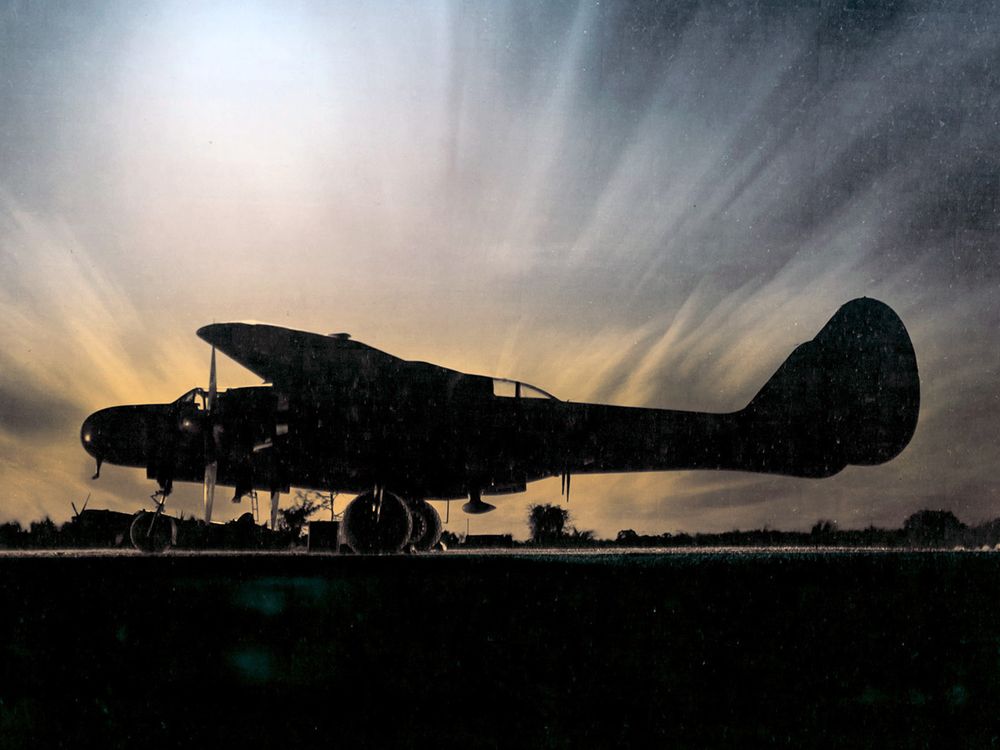 A military airplane painted in black sits on an airport ramp at sunset.