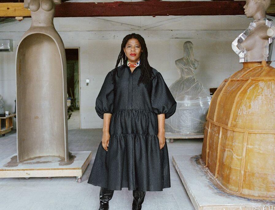 Simone Leigh, an African American woman, wears a voluminous black dress and stands in front of three of her sculptures, which are abstracted depictions of female figures on top of hoop skirts and jugs; Leigh faces the camera with a serious expression