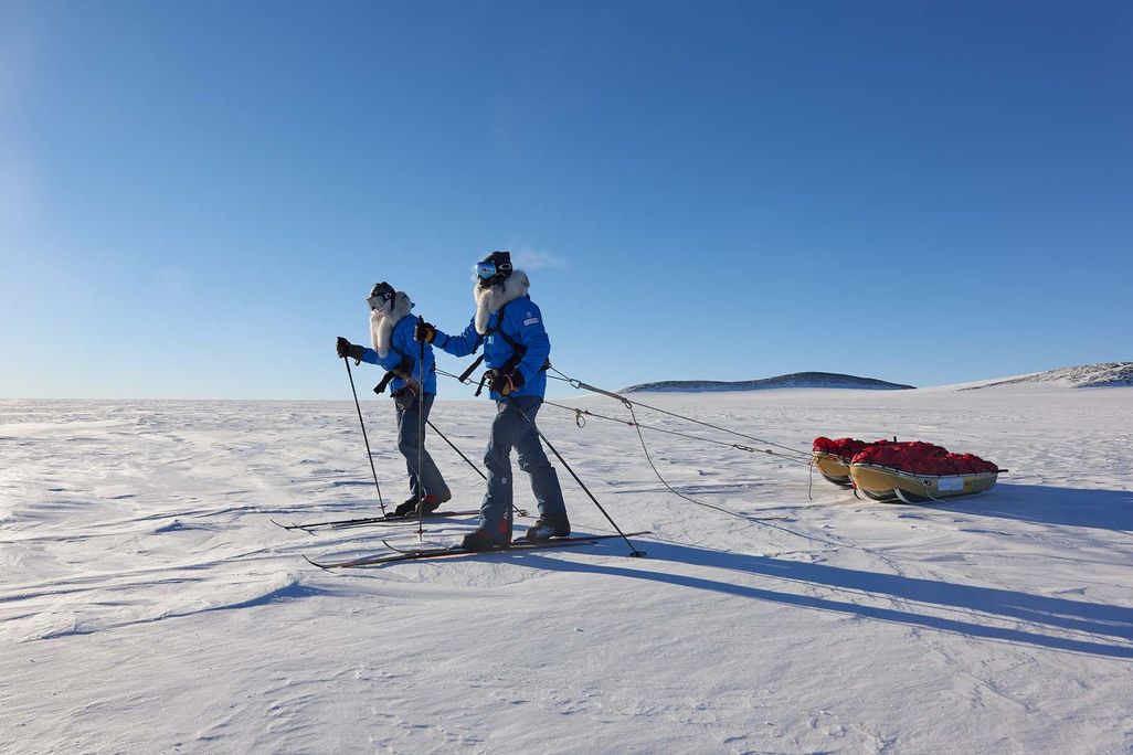 Two men in winter clothes and skis pull sleds across and frozen landscape