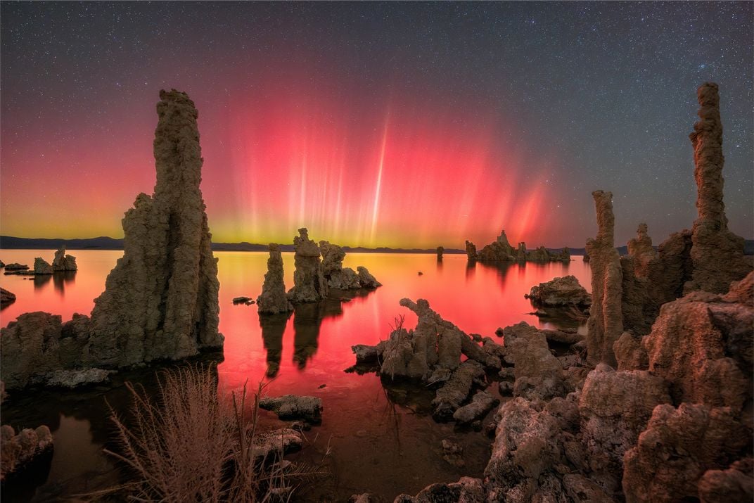 spires of glowing red, pink and yellow stretch up into the sky from the horizon and reflect into a lake, with tall rocky protrusions coming up across the landscape