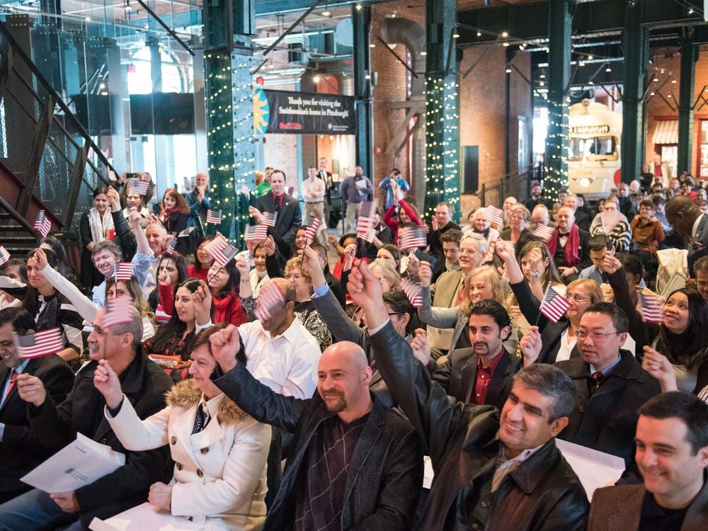 A group of people sit in chairs at the History Center raising mini American flags during a Citizenship ceremony