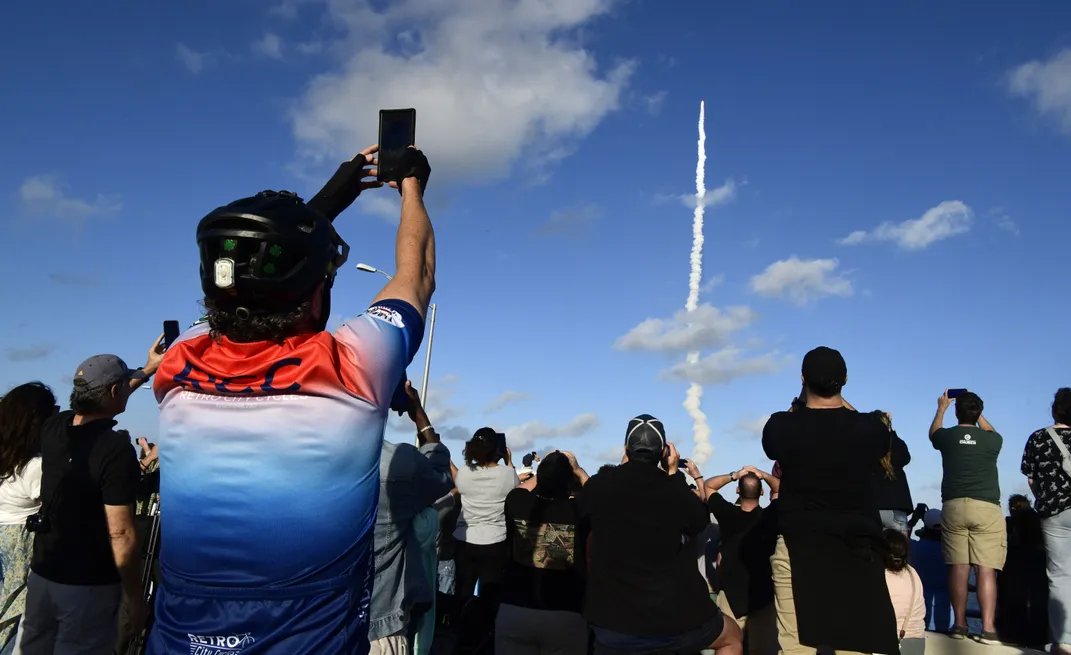 a person in biker's gear holds up a phone in front of the rocket launch