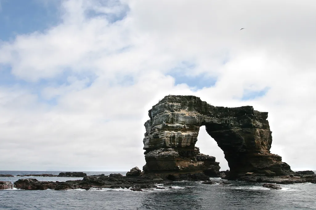 A photograph of Darwin's Arch in the Galàpagos Islands. The rock formation is two pillars attatched by a natural rock bridge. It stands itself in the ocean with a cloudy sky in the background.