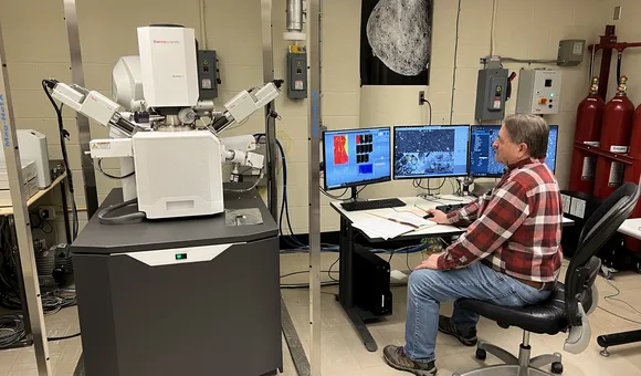 A man sits in front of a large, white machine and a row of computer screens in a lab setting.