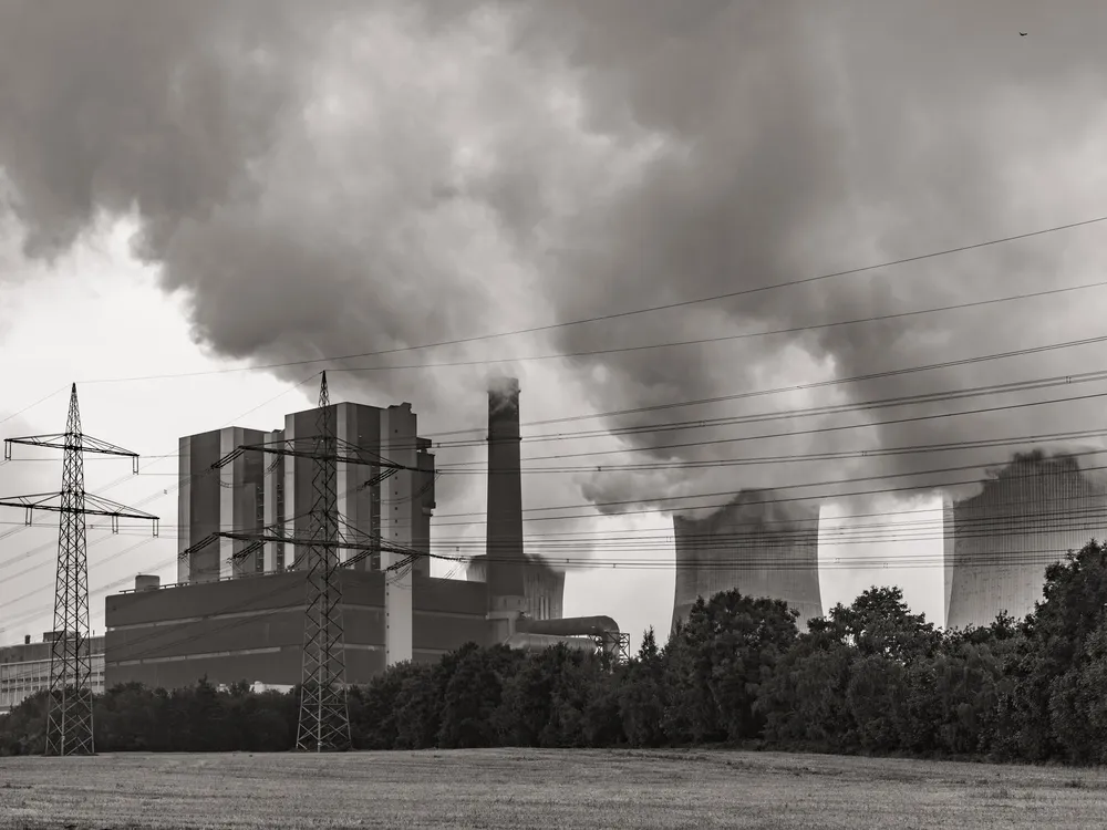 In black and white, power lines cross the foreground as a power plant billows smoke behind it.