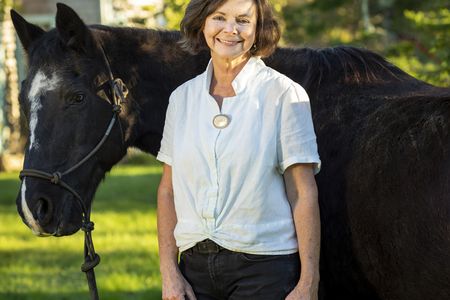 Image shows a woman wearing a white shirt standing with and holding the reins of a horse