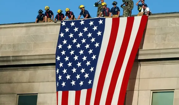 Several men stand on the roof of a large building, unfolding a red, white, and blue flag with a stars-and-stripes pattern over the side of the building.