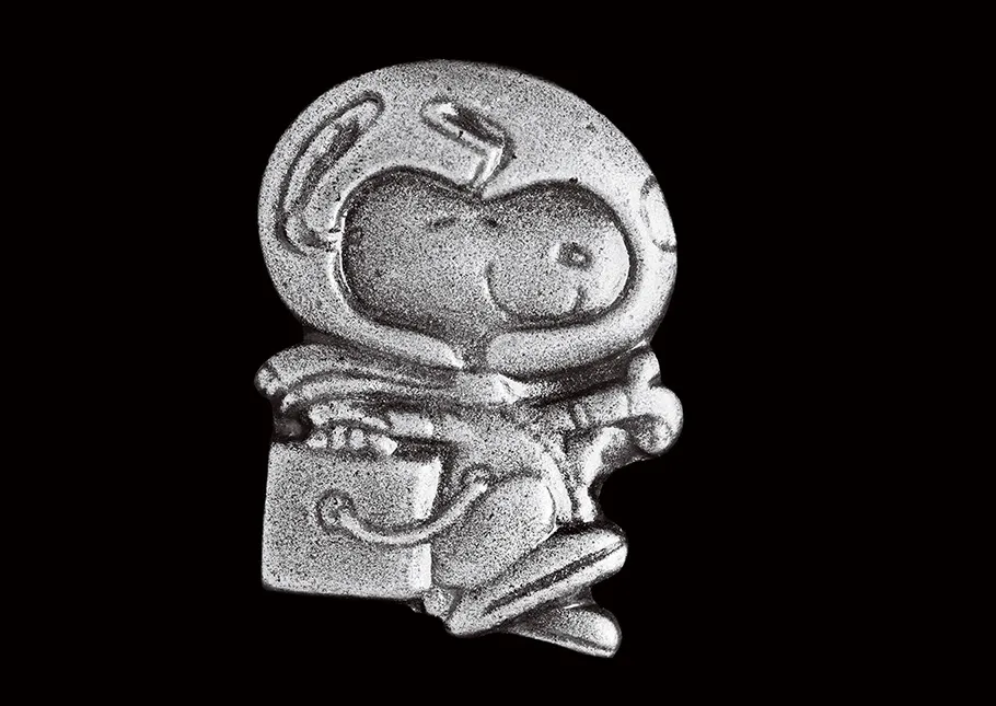 A small silver pin shows a smiling Snoopy, a cartoon dog, wearing a spacesuit with a large clear glass helmet, performing his iconic happy beagle dance.