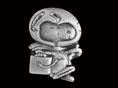 A small silver pin shows a smiling Snoopy, a cartoon dog, wearing a spacesuit with a large clear glass helmet, performing his iconic happy beagle dance.