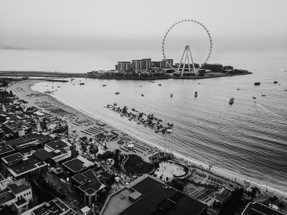 From the apex of the ferris wheel, the world unfolds in a perfect, medium-wide vista. This vantage point reveals the coastline's gentle embrace with the sea, framing the intimate meeting of sand and tide where people become tiny brushstrokes in a vast, romantic landscape.