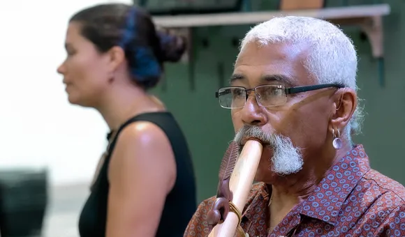 A man with short white hair, moustache, and glasses plays a wooden flute. Behind him, slightly out of focus, is a young woman with long dark hair in a bun and a black tank top.