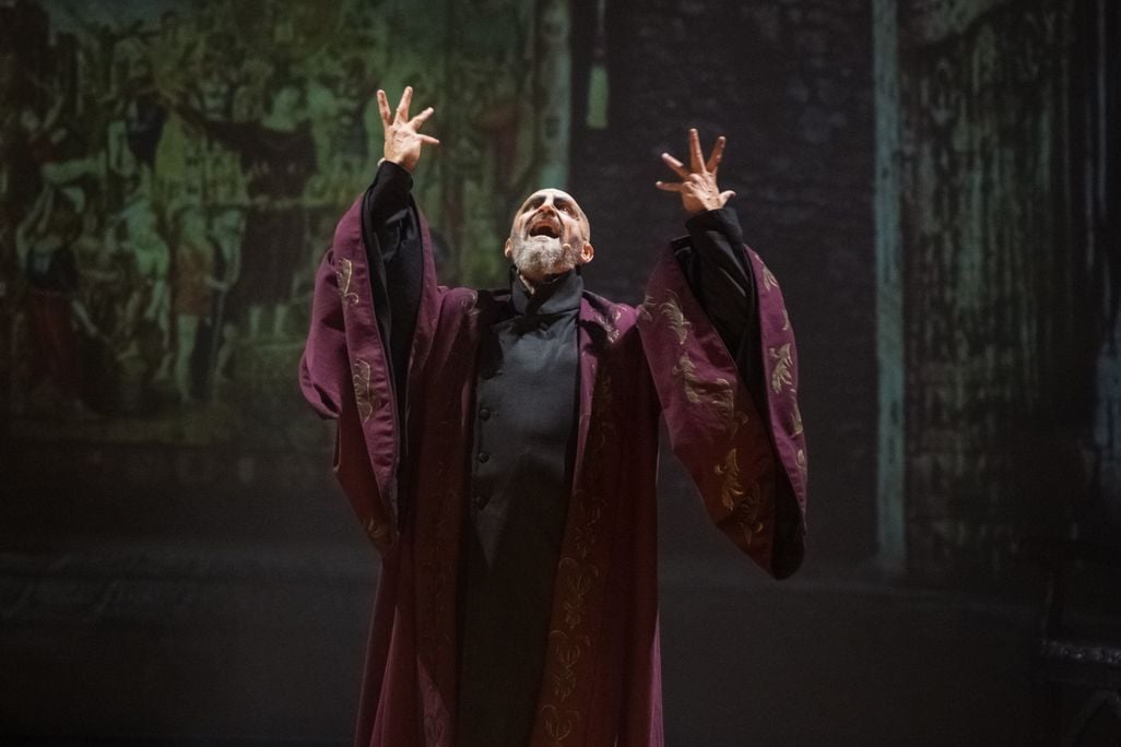 Man wearing a burgundy robe sings with arms outstretched on stage