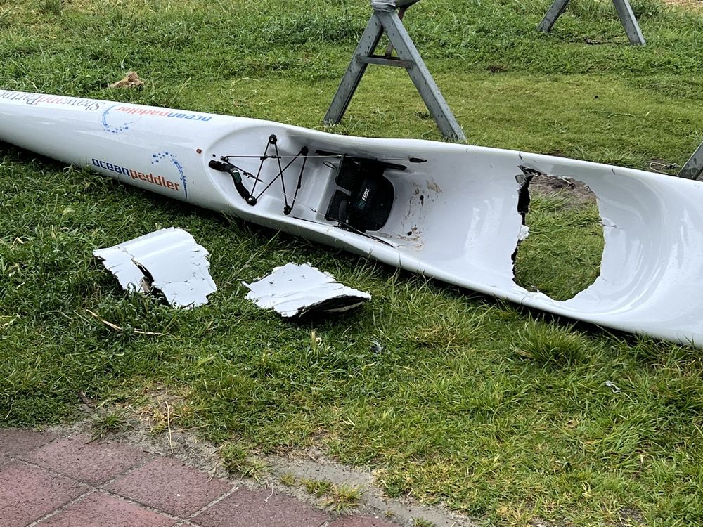 A white surf ski with a hole torn into it on the grass in front of a beach