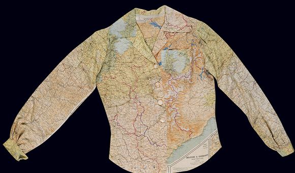 A ladies long-sleeve blouse fastened by four plastic white buttons hangs against a dark background. The blouse features a multi-color map pattern printed on a white silk background.