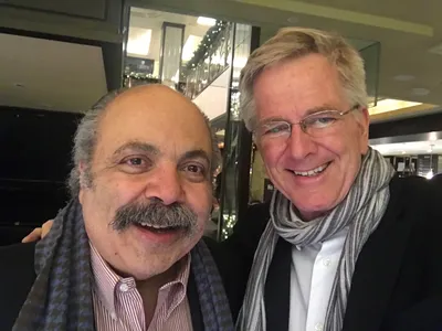 Two men stand, one wearing glasses and a scarf and the other with a mustache, smile for the camera.