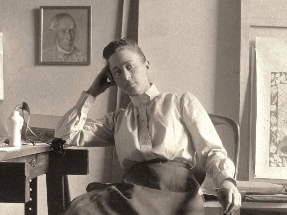 Hilma af Klint, a white woman in a white shirt and a long black skirt, leans her hand on one elbow and sits at a desk in her studio