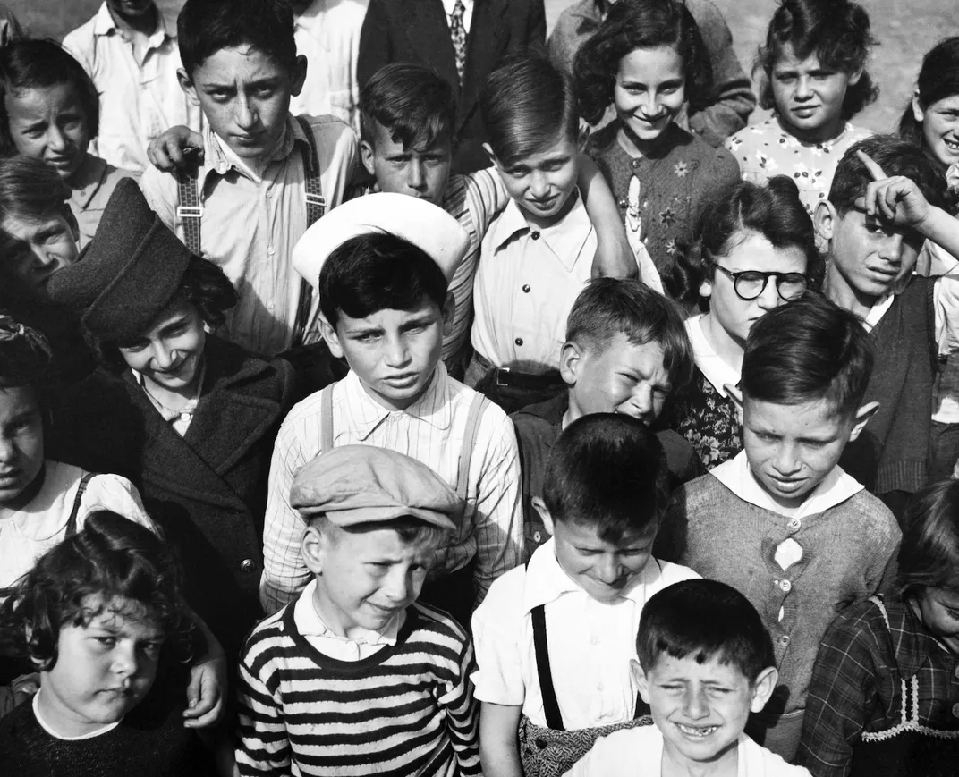 A black and white image of many young children looking upward at the camera, many squinting as though bothered by the sun