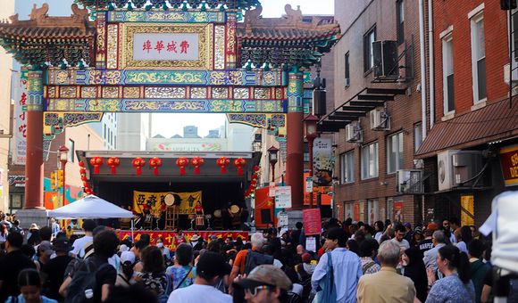 A crowd gathers on a street, where a stage is set up under an elaborate, decorative Chinese-style archway.