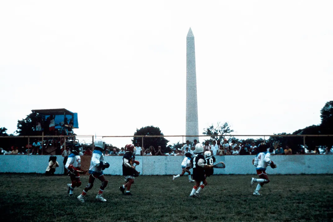 A group of kids in uniforms play lacrosse on the grass of the National Mall, with the Washington Monument in the background.