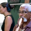 A man with short white hair, moustache, and glasses plays a wooden flute. Behind him, slightly out of focus, is a young woman with long dark hair in a bun and a black tank top.