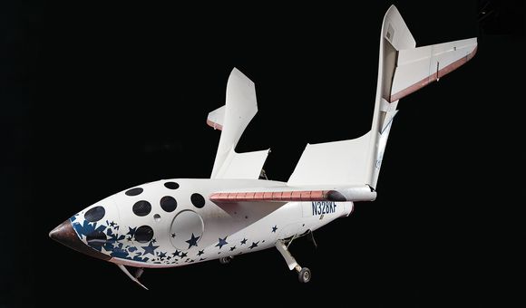 A white spacecraft against a black background. The fuselage is spangled with dark blue stars and black circles. A large wing is swiveled up away from the fuselage. The tail of the spacecraft is marked by this identifying number: N328KF.