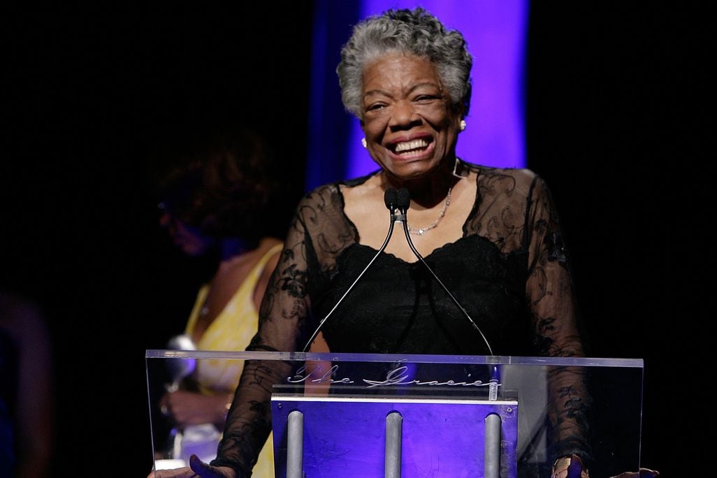 Angelou in front of a podium on a stage in a black formal dress, in front of a purple backdrop, smiling