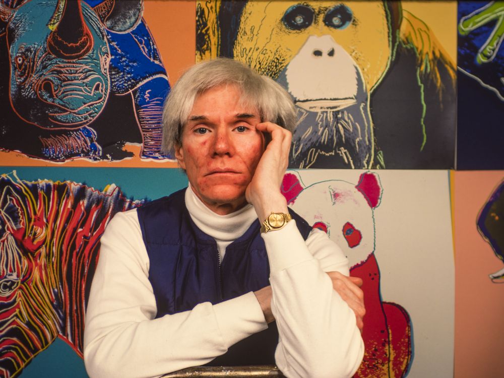 A color image of Warhol, wearing a floppy white wig, a white turtleneck and blue vest, poses with his head on his hand in front of screen printed works featuring rhinos, zebras, and pandas