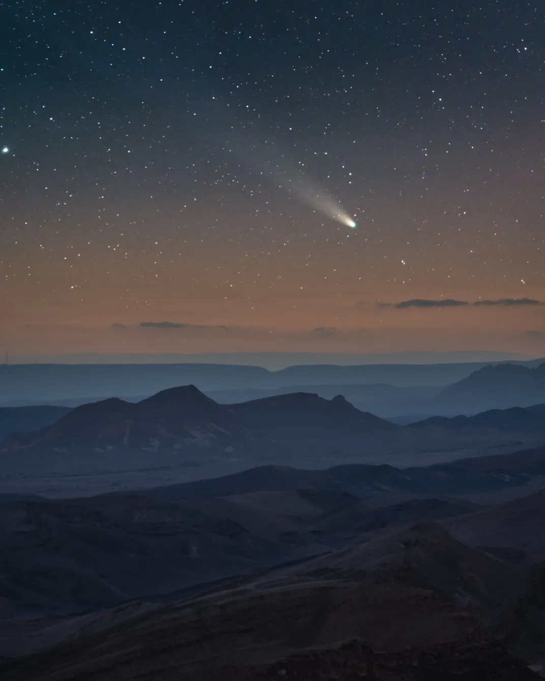 a comet streaks white above mountains of sand in a desert. the sky appears orange and stars are flecks of light