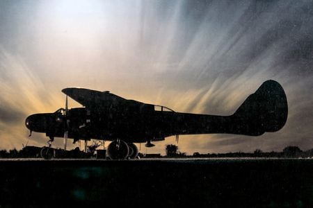 A military airplane painted in black sits on an airport ramp at sunset.