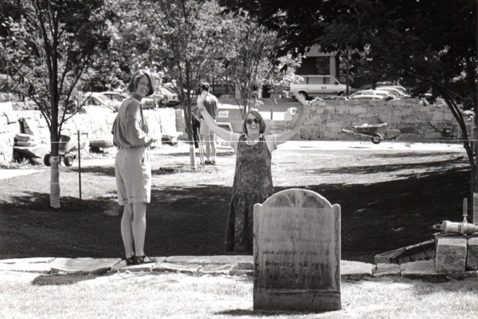 Two women smile from a construction site outdoors. A gravestone is in the foreground.