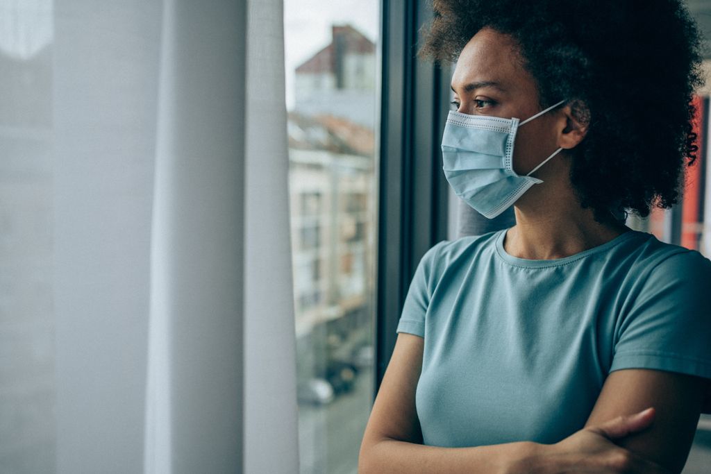 A Black woman with a face masks looks out of a window
